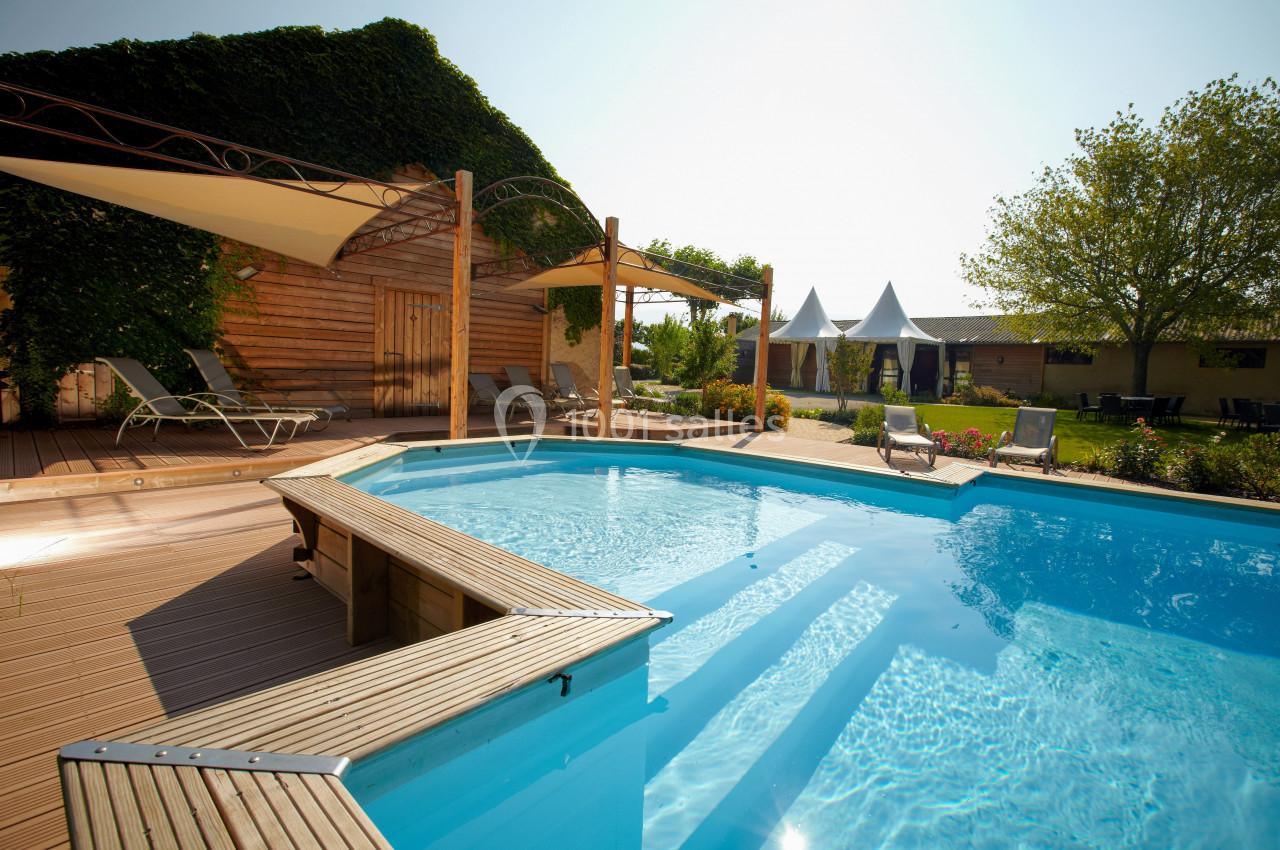 Piscine extérieure entourée de terrasses en bois, transats et espaces verts sous un ciel dégagé.
