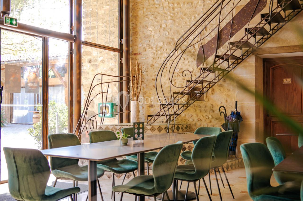 Salle lumineuse avec murs en pierre, escalier en fer forgé, grandes baies vitrées et chaises vertes autour de tables.