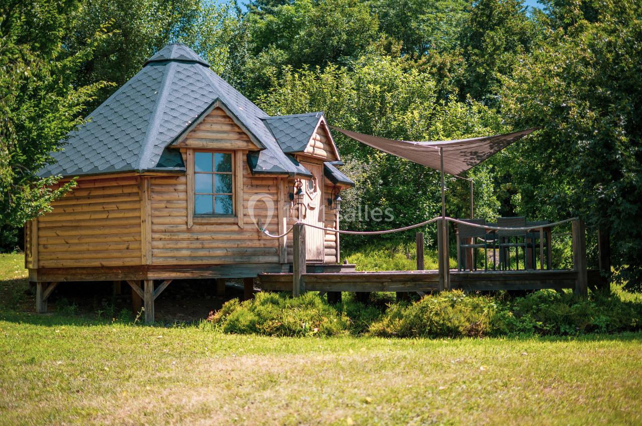 Cabane en bois avec toit en ardoise, terrasse en bois et voile d'ombrage, entourée de verdure.