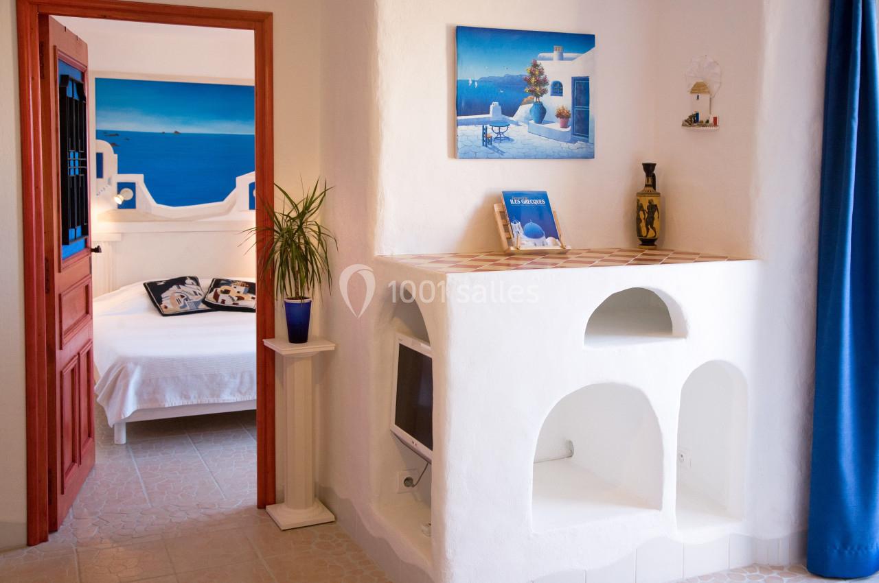 Intérieur lumineux d'une chambre méditerranéenne avec murs blancs, décor marin et vue sur un lit à travers une porte ouverte.