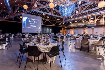 Un artiste sur échasses interagit avec une femme dans un espace intérieur éclairé, avec des spectateurs en arrière-plan.