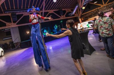Un artiste sur échasses interagit avec une femme dans un espace intérieur éclairé, avec des spectateurs en arrière-plan.