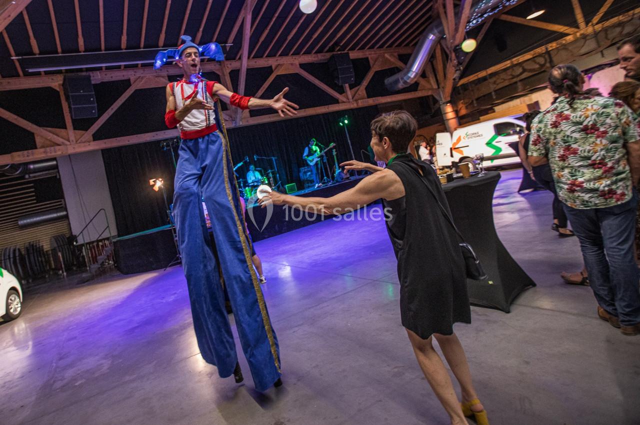 Un artiste sur échasses interagit avec une femme dans un espace intérieur éclairé, avec des spectateurs en arrière-plan.