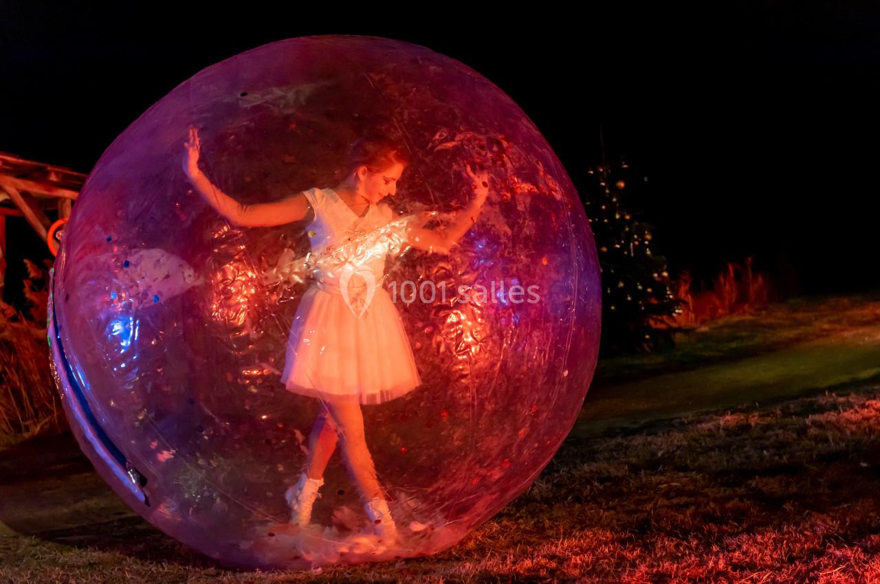 Femme en robe blanche dans une grande bulle transparente, éclairée par des lumières colorées dans un environnement nocturne.