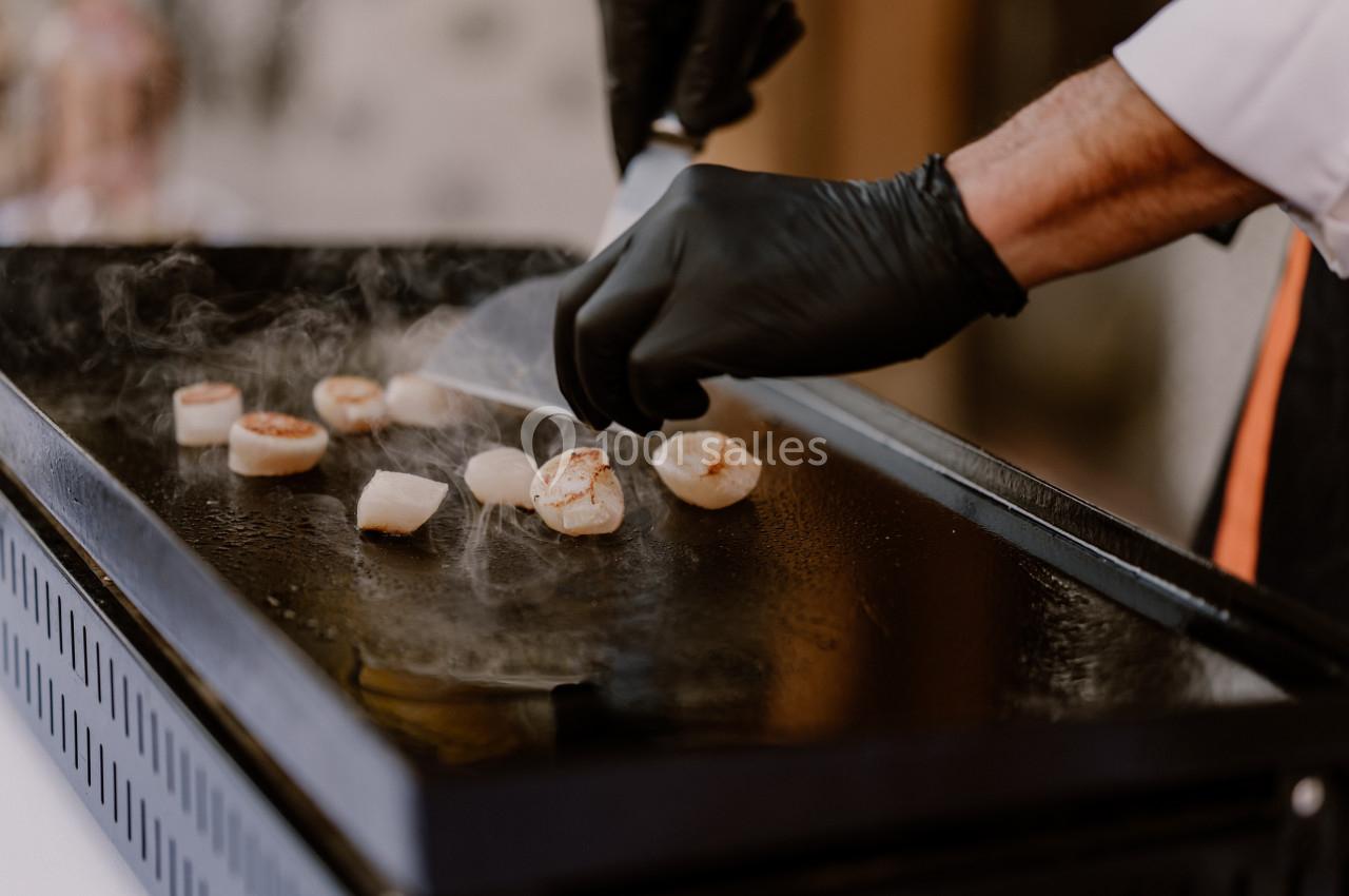 Un chef fait griller des noix de Saint-Jacques sur une plaque chauffante, portant des gants noirs.