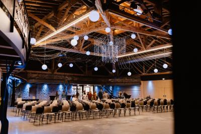 Un artiste sur échasses interagit avec une femme dans un espace intérieur éclairé, avec des spectateurs en arrière-plan.