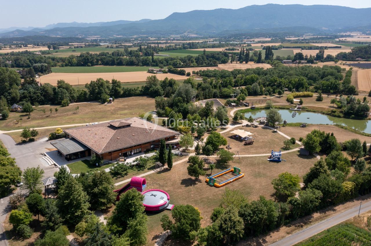 Vue aérienne d'un parc avec bâtiments, structures gonflables, étang et paysages vallonnés en arrière-plan.