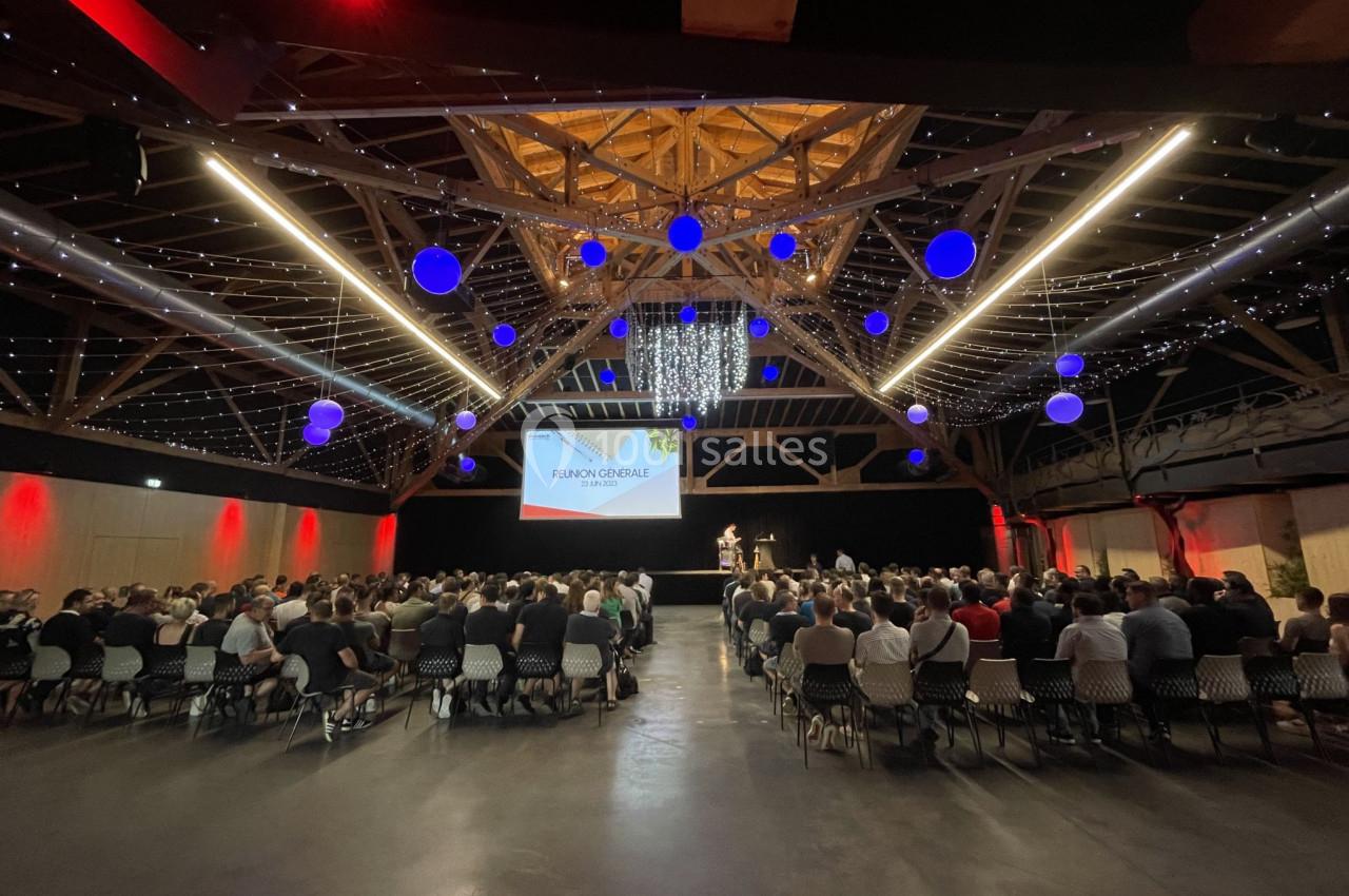 Salle comble avec des participants assis face à une scène, sous une charpente en bois illuminée et des décorations…