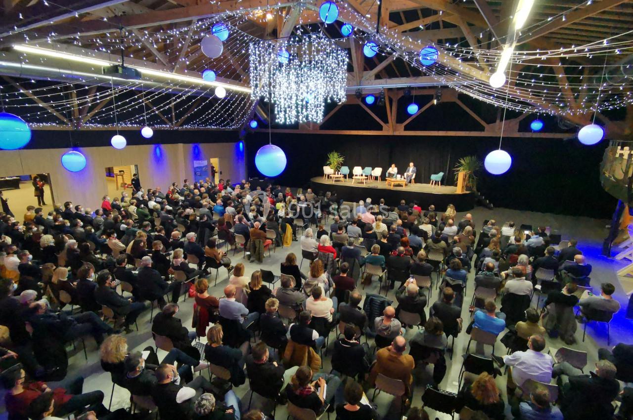 Salle comble avec un public assis, éclairée par des guirlandes lumineuses et des décorations suspendues, lors d'un événement.