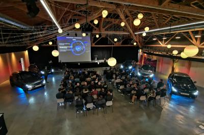 Un artiste sur échasses interagit avec une femme dans un espace intérieur éclairé, avec des spectateurs en arrière-plan.