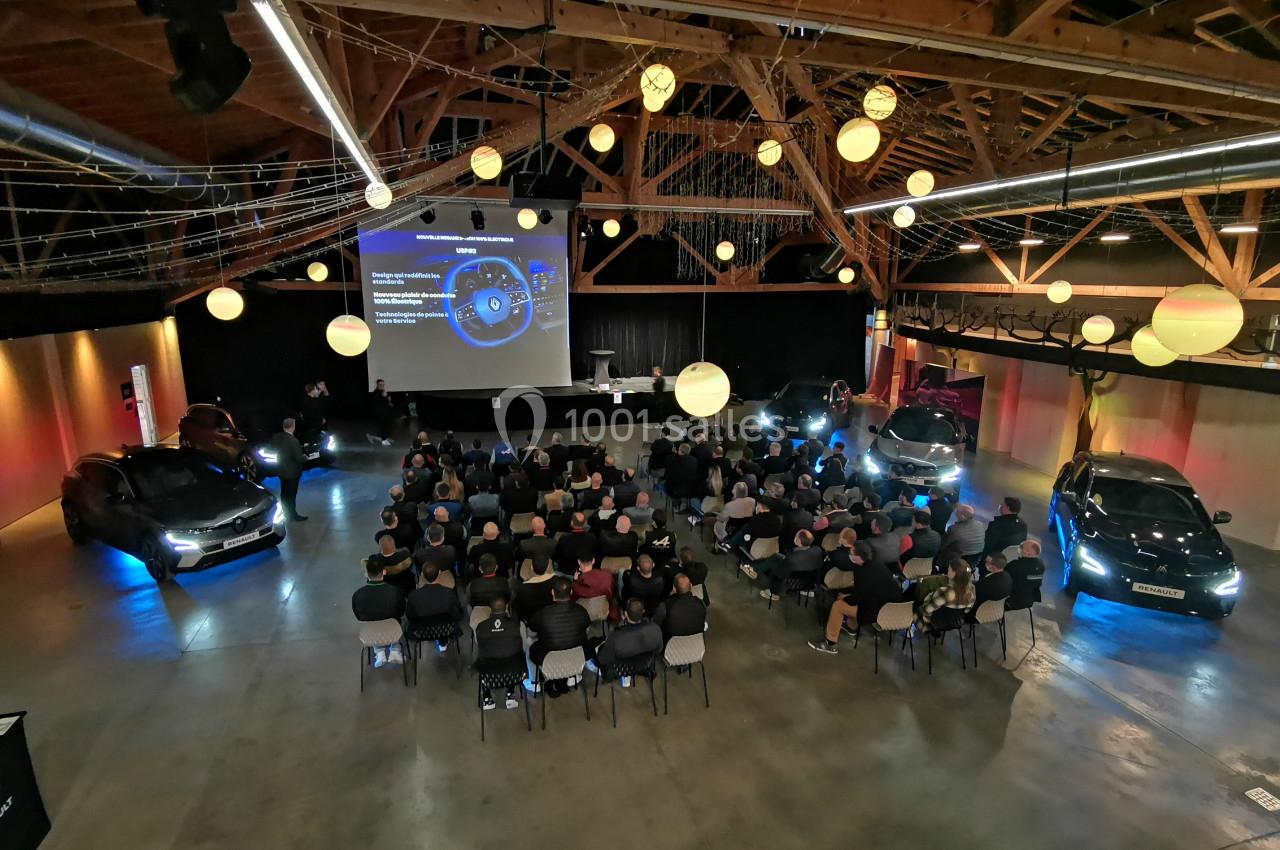 Salle de conférence avec des participants assis, des voitures exposées et une présentation projetée sur écran.