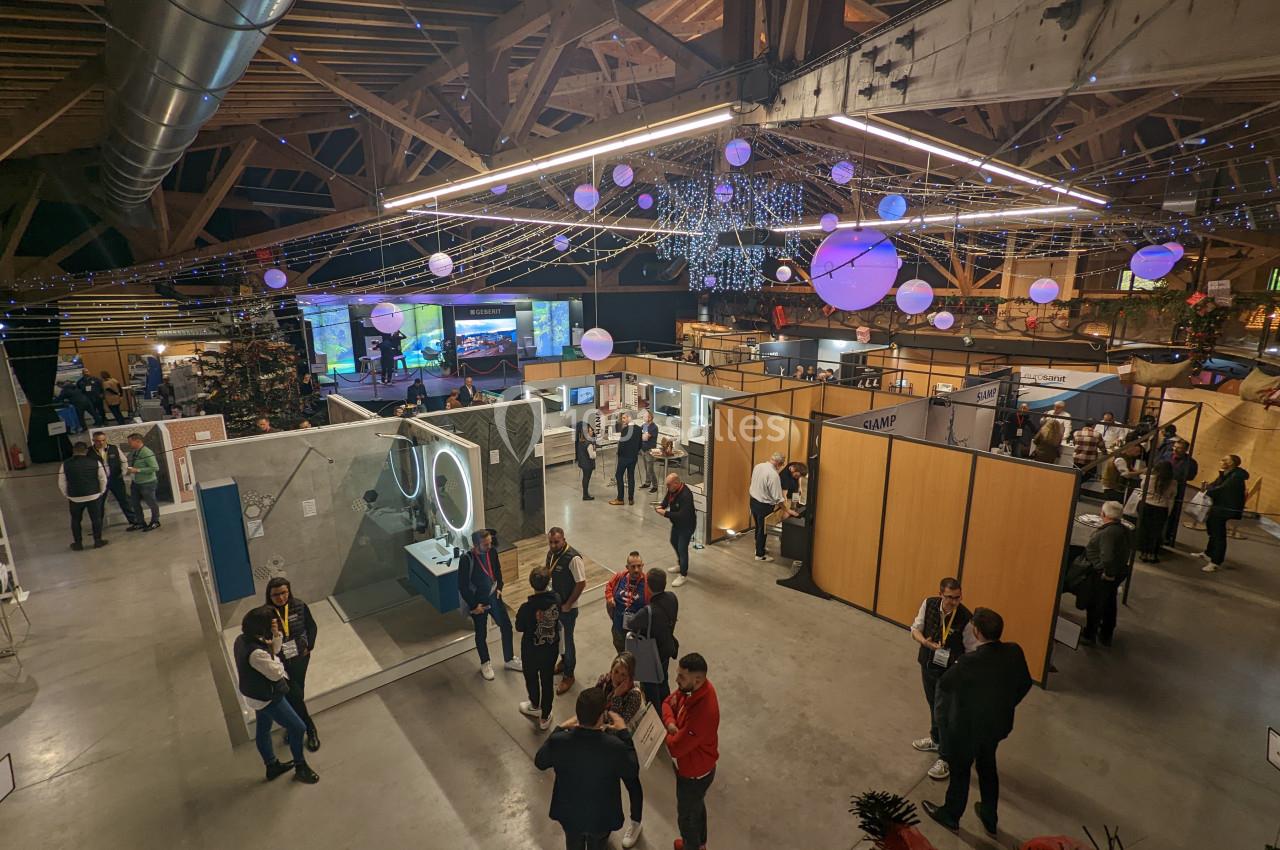 Vue d'un espace d'exposition intérieur avec des stands, des visiteurs et une décoration lumineuse suspendue.