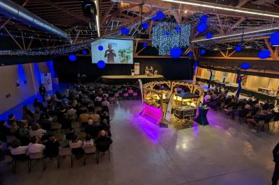Un artiste sur échasses interagit avec une femme dans un espace intérieur éclairé, avec des spectateurs en arrière-plan.