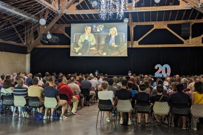 Un artiste sur échasses interagit avec une femme dans un espace intérieur éclairé, avec des spectateurs en arrière-plan.