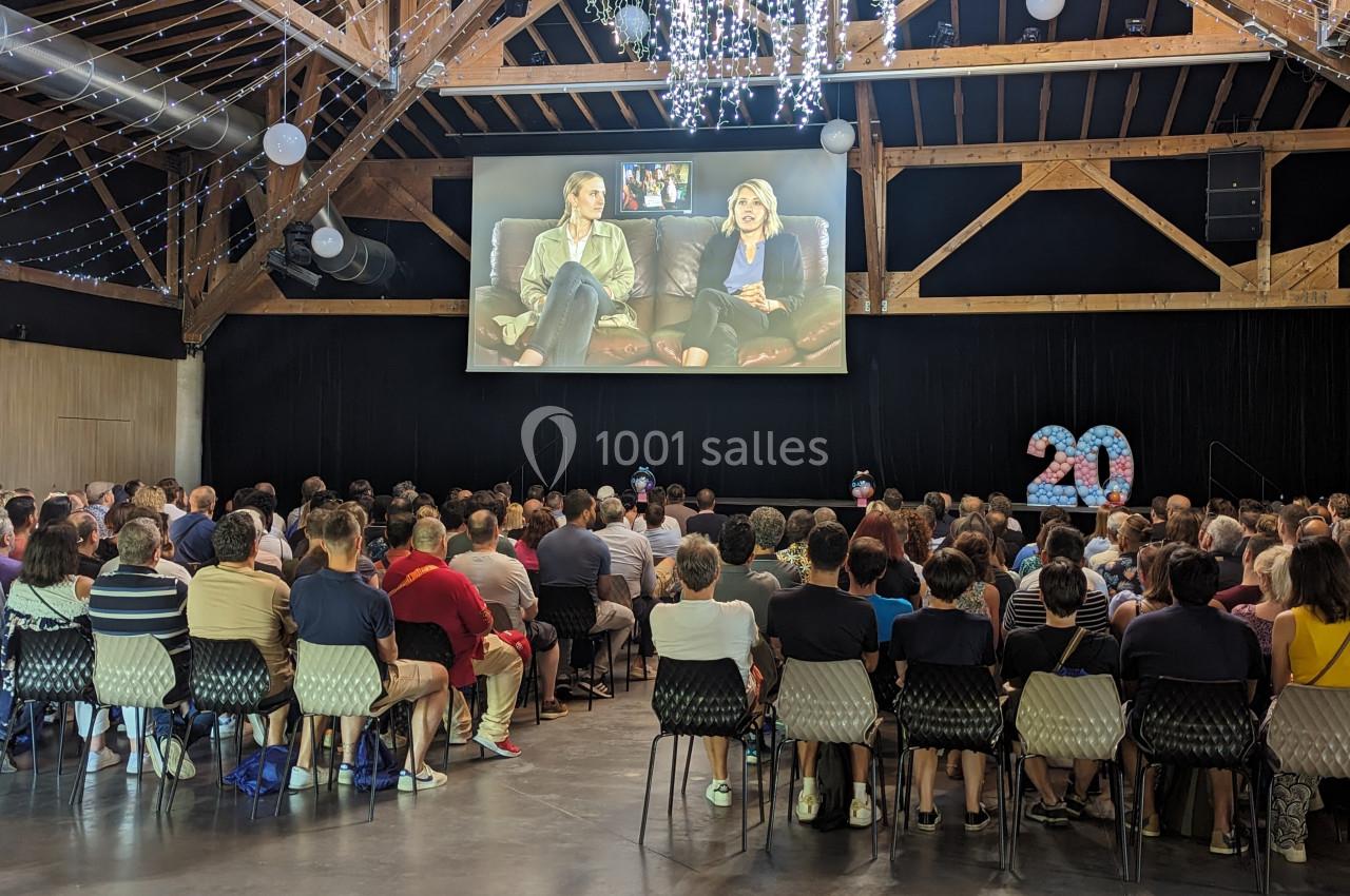 Un public assis regarde une projection sur grand écran dans une salle avec des poutres apparentes et des décorations…