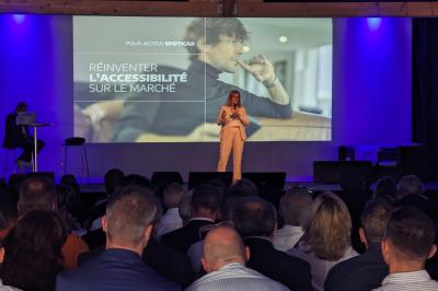 Un artiste sur échasses interagit avec une femme dans un espace intérieur éclairé, avec des spectateurs en arrière-plan.