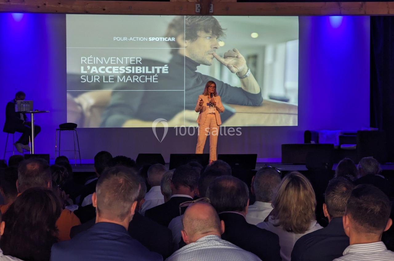 Une femme présente sur scène devant un public, avec une projection affichant ’Réinventer l'accessibilité sur le marché’.