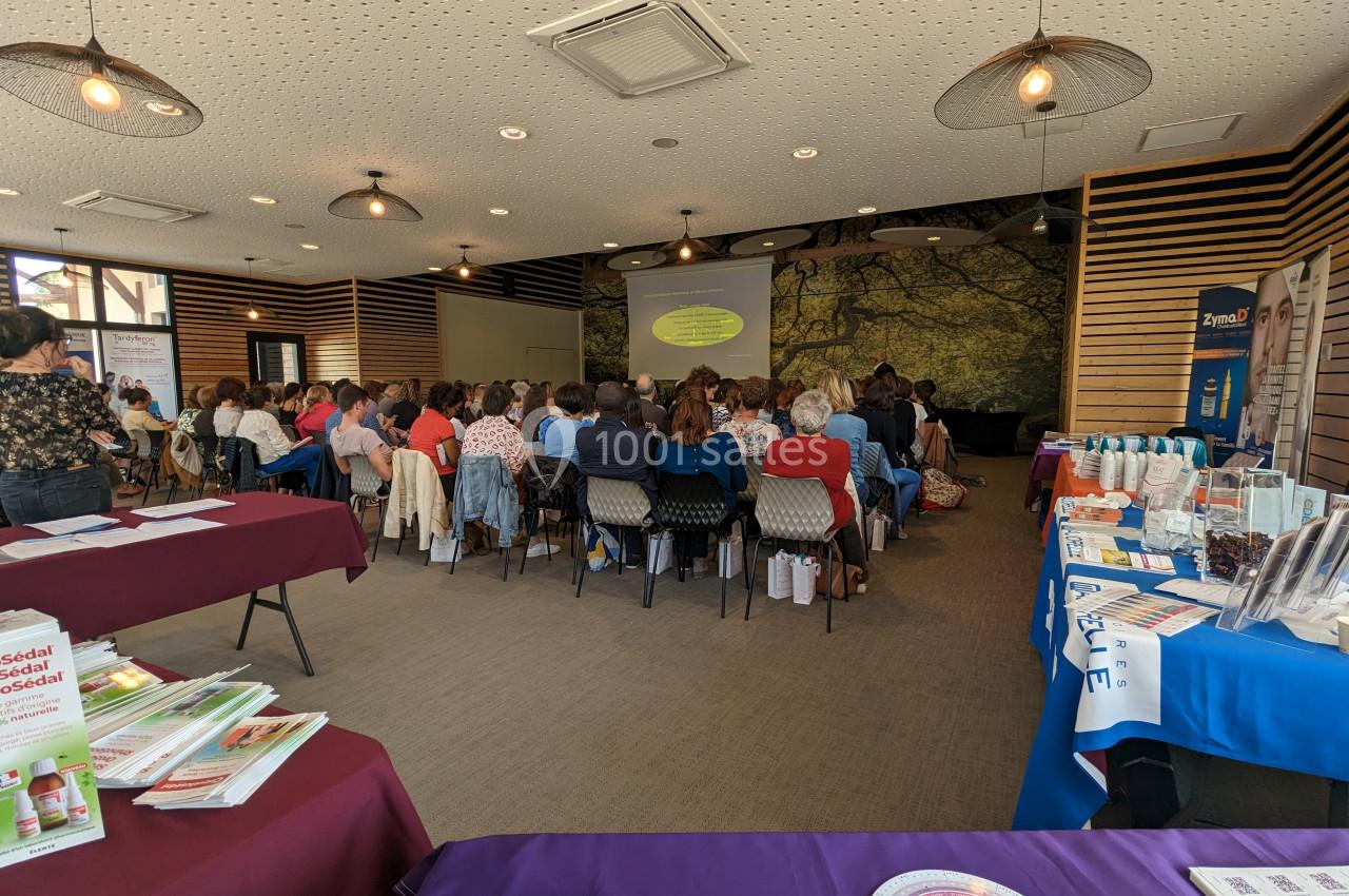 Salle de conférence avec un public assis, regardant une présentation projetée, entourée de stands d'information.