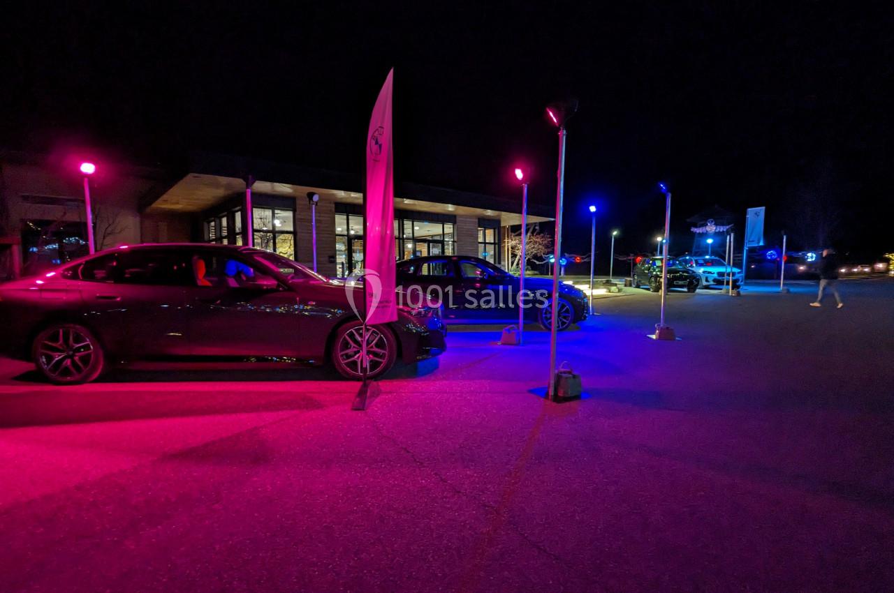 Voitures exposées de nuit sur un parking éclairé par des lumières colorées, avec un bâtiment en arrière-plan.