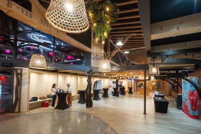 Un artiste sur échasses interagit avec une femme dans un espace intérieur éclairé, avec des spectateurs en arrière-plan.