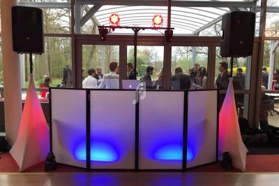 Pupitre de DJ éclairé en bleu et rouge, entouré de haut-parleurs, avec des invités discutant en arrière-plan.