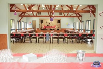 Salle décorée pour un événement avec tables alignées, nappes rouges et blanches, ballons suspendus et espace pour la musique.