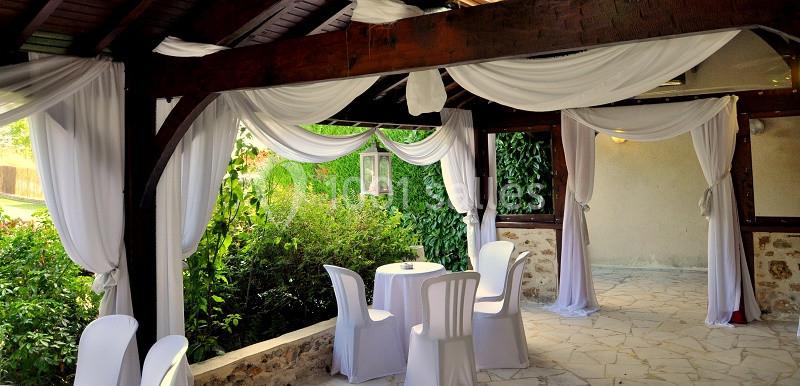 Terrasse couverte décorée de voilages blancs, avec des chaises et une table ronde, entourée de verdure.