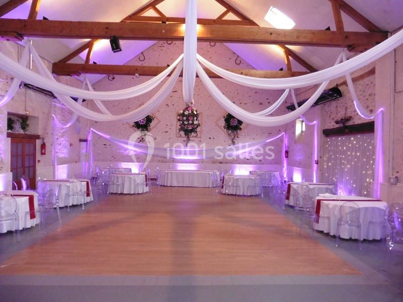 Salle de réception décorée avec des drapés blancs suspendus, éclairage violet et tables dressées autour d'une piste en bois.