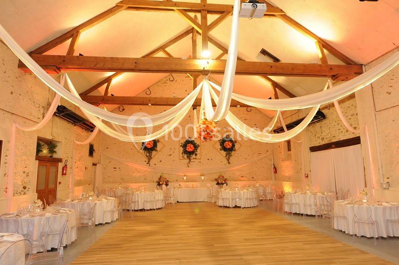 Salle de réception décorée avec des guirlandes blanches suspendues, des tables rondes dressées et des fleurs colorées.