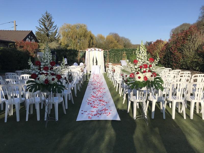 Allée centrale décorée de pétales de rose menant à une arche fleurie, entourée de chaises blanches en extérieur.