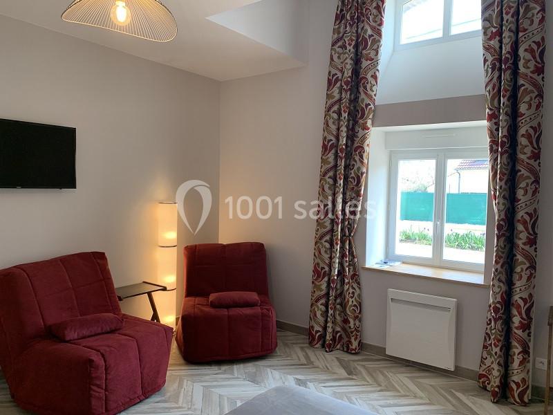 Deux fauteuils rouges dans un salon lumineux avec une fenêtre, des rideaux à motifs et un téléviseur mural.