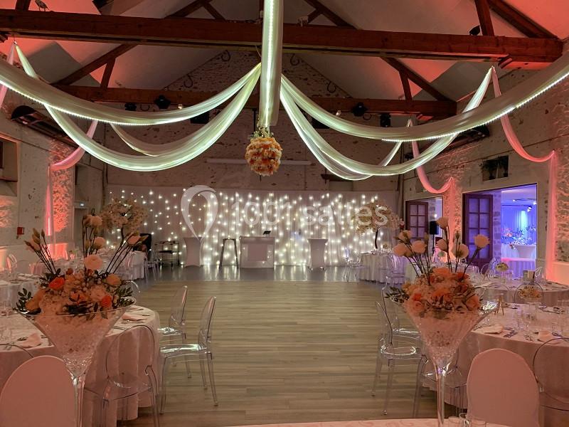 Salle de réception décorée avec des guirlandes lumineuses, des drapés suspendus et des compositions florales sur les tables.