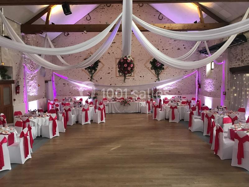 Salle de réception décorée pour un événement, avec des tables dressées, des nappes blanches et des nœuds roses.