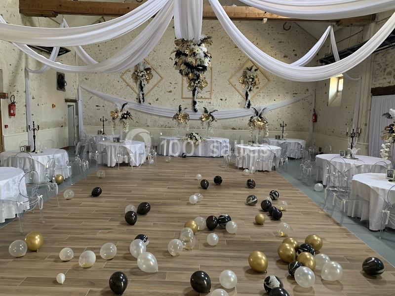 Salle de réception décorée avec des drapés blancs, des tables dressées et des ballons noirs, blancs et dorés au sol.