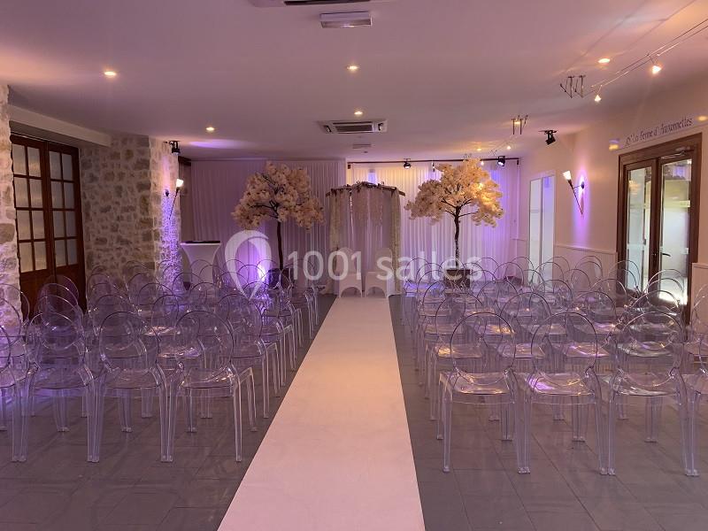 Salle décorée pour une cérémonie avec chaises transparentes alignées, allée centrale blanche et arbres artificiels fleuris.