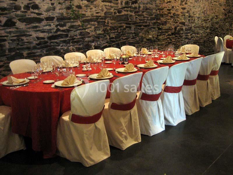 Table dressée avec une nappe rouge, des assiettes, des verres et des serviettes pliées, entourée de chaises blanches.