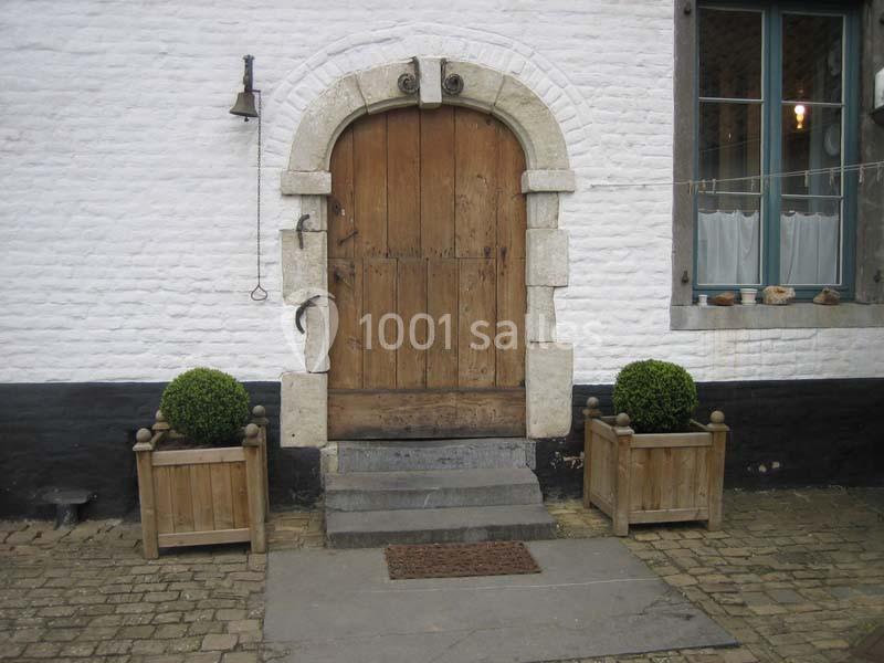 Location salle Court-Saint-Étienne (Brabant wallon) - Ferme de Beaurieux #4 Porte en bois ancienne avec encadrement en pierre, flanquée de deux bacs à plantes sur un sol pavé.