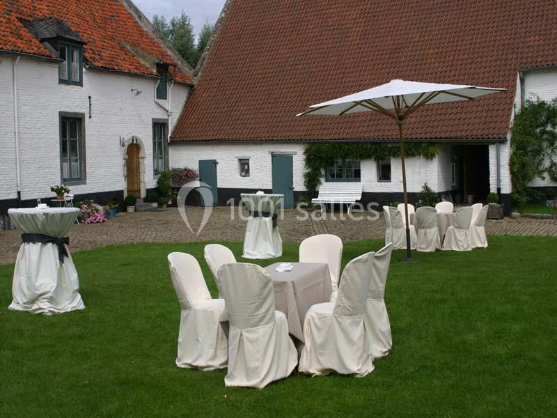 Location salle Court-Saint-Étienne (Brabant wallon) - Ferme de Beaurieux #9 Chaises et tables habillées de housses blanches disposées sur une pelouse devant des bâtiments à toits en tuiles.