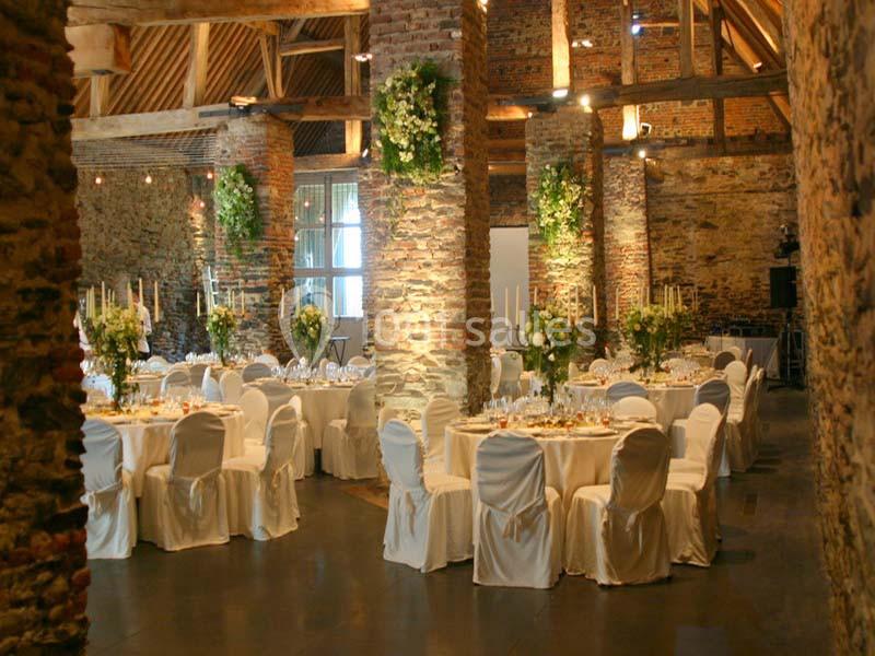 Salle de réception en pierre avec tables rondes dressées, chaises blanches et décorations florales suspendues.