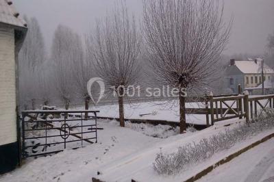 Paysage hivernal avec arbres dénudés, sol enneigé, clôture en bois et maisons en arrière-plan sous un ciel gris.