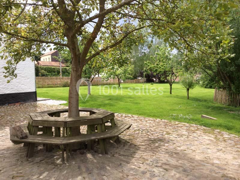 Banc en bois circulaire autour d'un arbre, sur une cour pavée, avec un jardin verdoyant en arrière-plan.