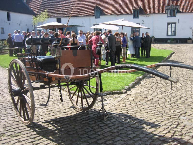 Une calèche ancienne en bois est stationnée sur des pavés devant un groupe de personnes réunies dans une cour.