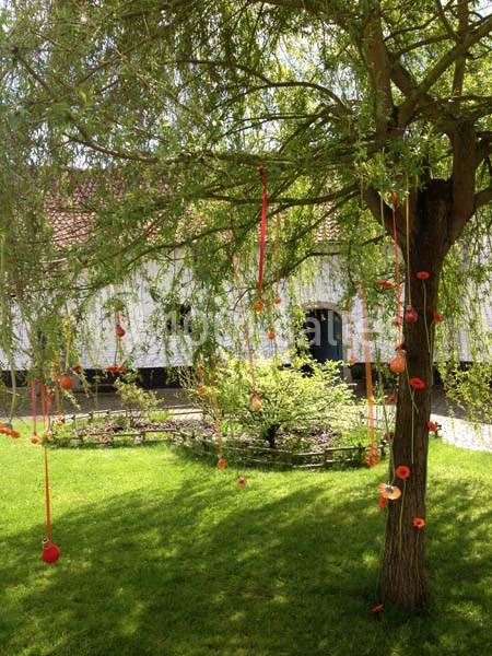 Arbre décoré de rubans rouges et d'objets suspendus dans un jardin verdoyant devant une maison blanche.