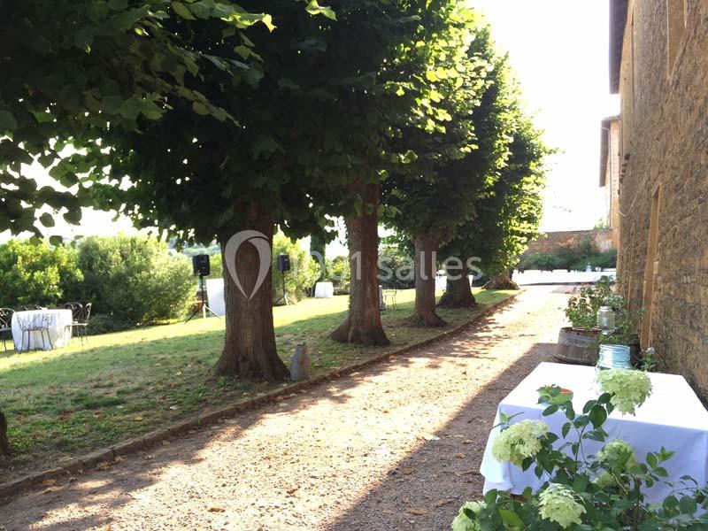 Allée bordée d'arbres avec des tables dressées en arrière-plan dans un jardin ensoleillé.