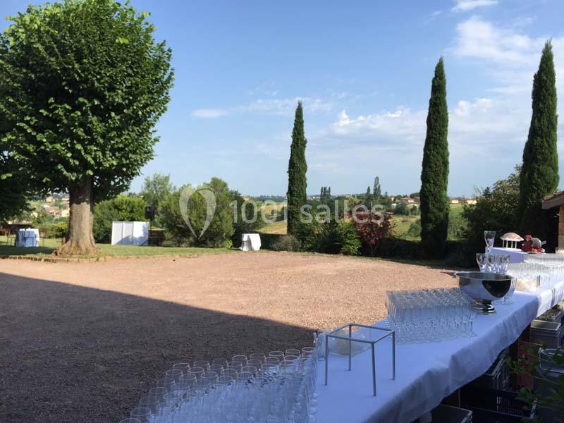 Tables dressées avec des verres et des coupes en extérieur, devant un paysage rural avec arbres et cyprès.