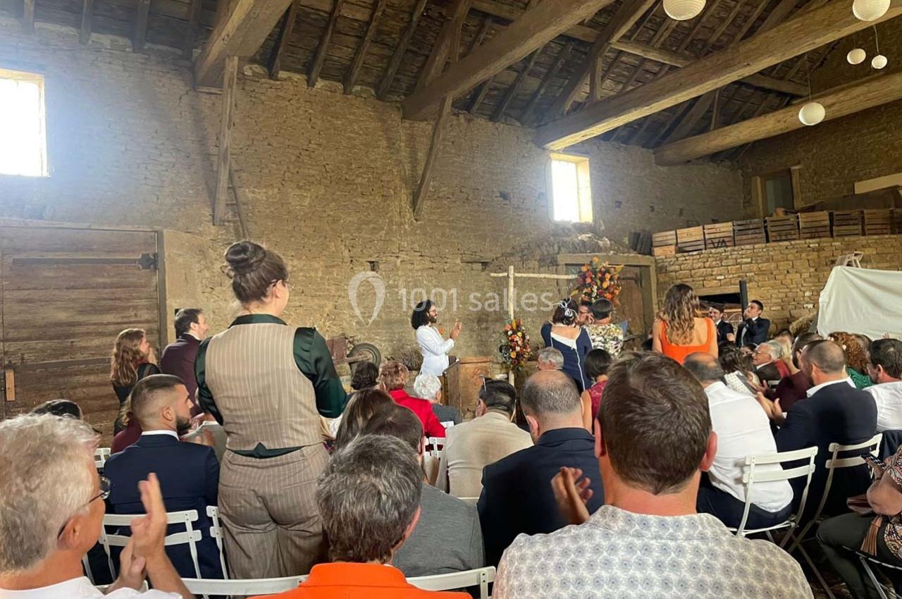 Des personnes assistent à une cérémonie dans une grange en pierre avec une décoration florale et des chaises blanches.