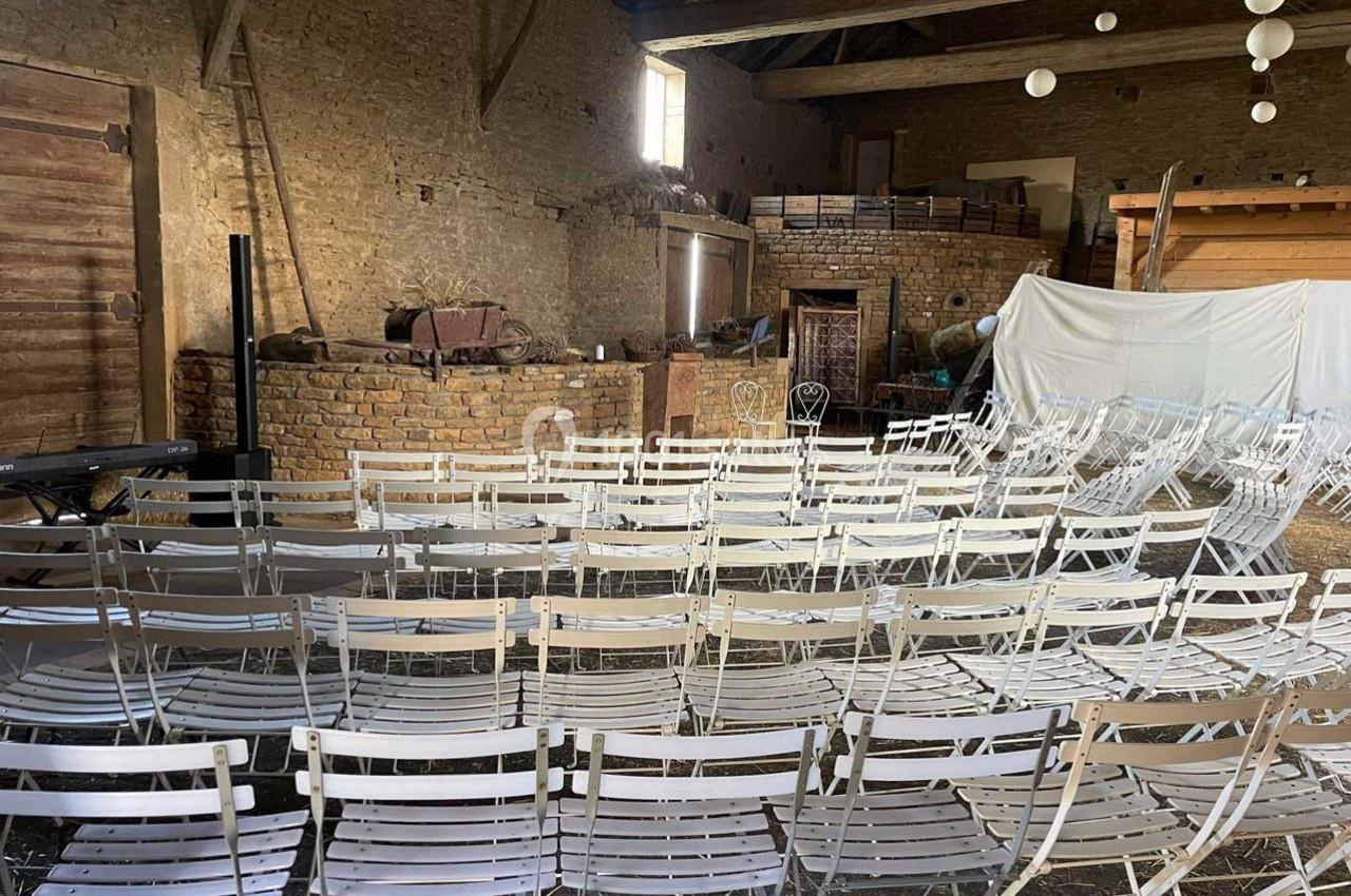 Chaises blanches pliantes alignées dans une grange en pierre, face à une scène improvisée avec des éléments rustiques.