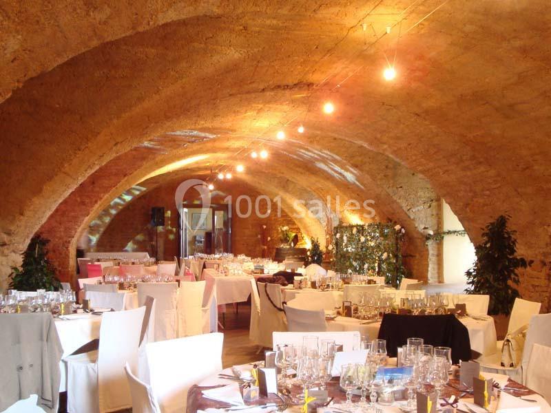Salle voûtée en pierre aménagée pour un événement, avec tables dressées, chaises blanches et éclairage chaleureux.