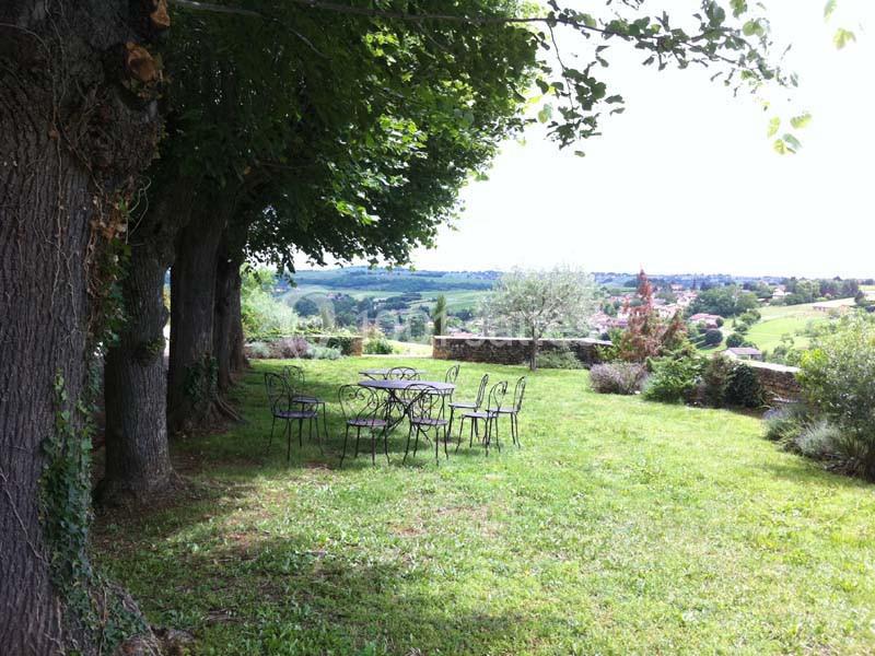 Table et chaises en fer forgé sur une pelouse ombragée, avec vue sur une campagne vallonnée et des arbres.
