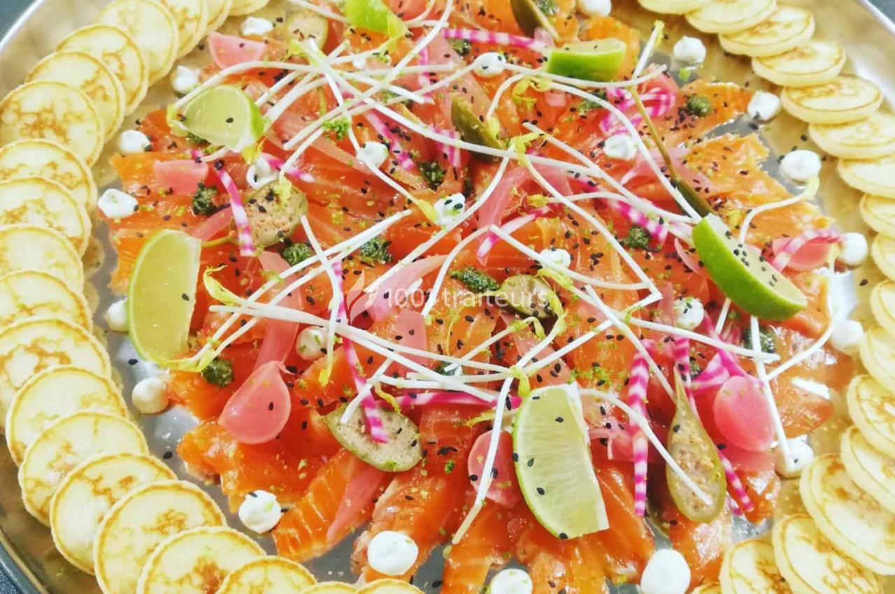 Assiette de saumon mariné avec tranches de citron vert, légumes marinés, herbes fraîches et mini-blinis en bordure.
